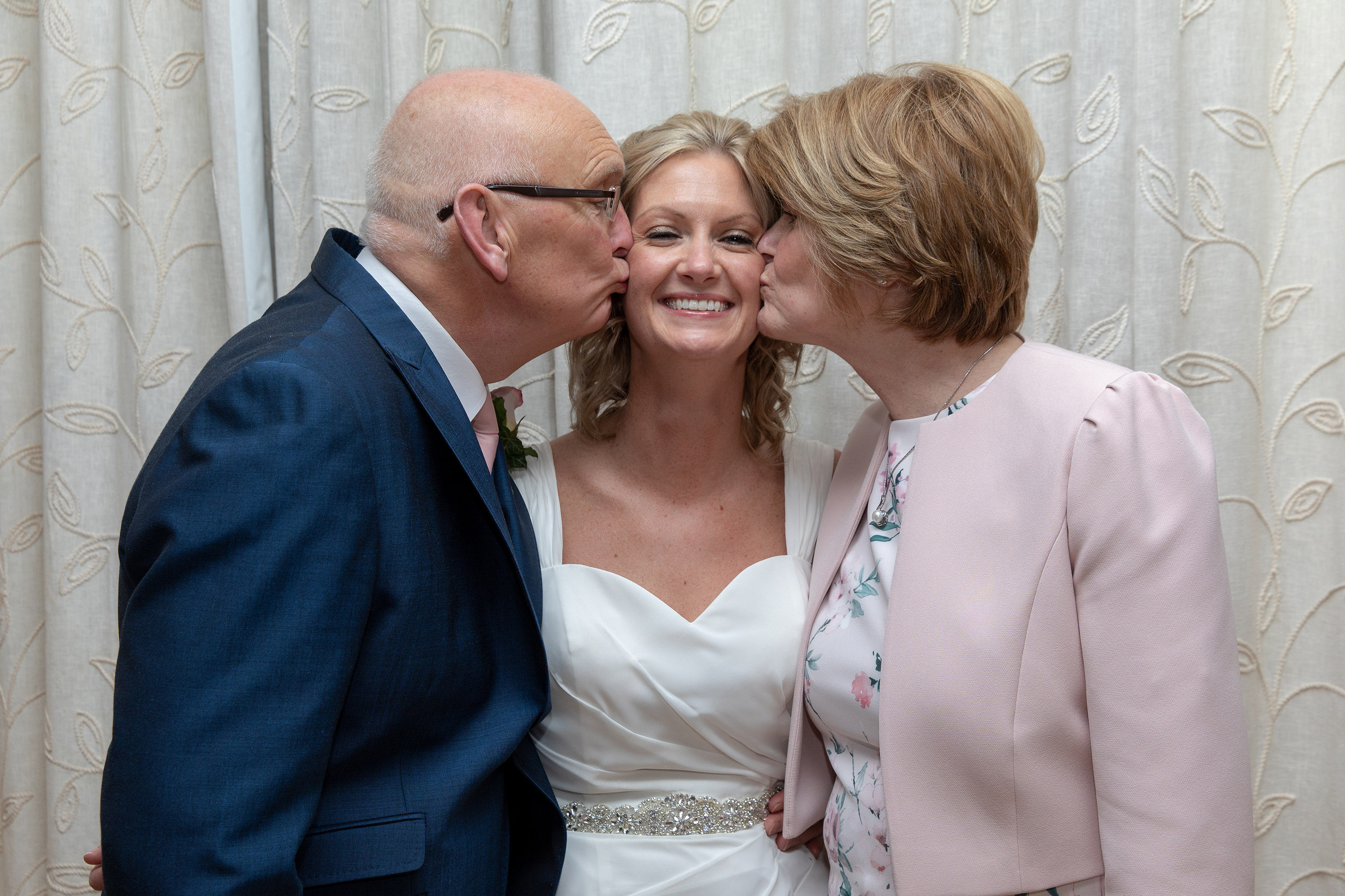 mother and father of the bride and groom give her a kiss